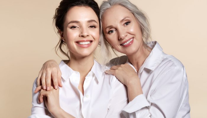 Two beautiful woman in white shirt hug face forward portrait. Studio headshot of toothy smiling grandmother and granddaughter, senior mother and daughter, elderly and young sister. Happy family