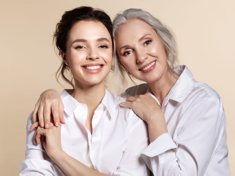 Two beautiful woman in white shirt hug face forward portrait. Studio headshot of toothy smiling grandmother and granddaughter, senior mother and daughter, elderly and young sister. Happy family